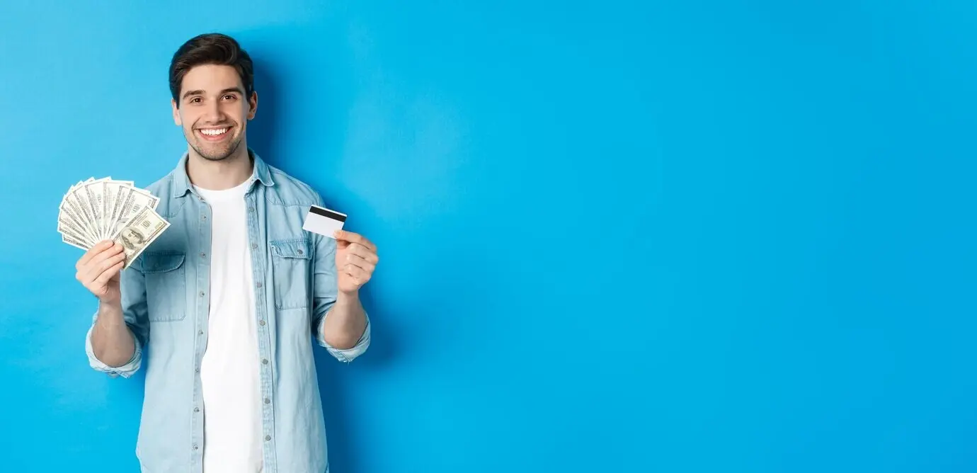 Hombre joven sonriente mostrando dólares en efectivo y una tarjeta de crédito, de pie frente a un fondo azul.