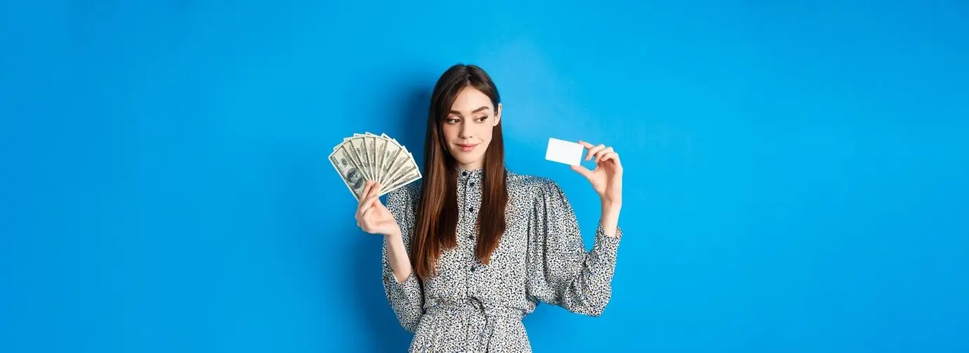 Chica hermosa de compras con vestido, mirando hacia un lado y sonriendo pensativa, mostrando dinero y una tarjeta de crédito de plástico.