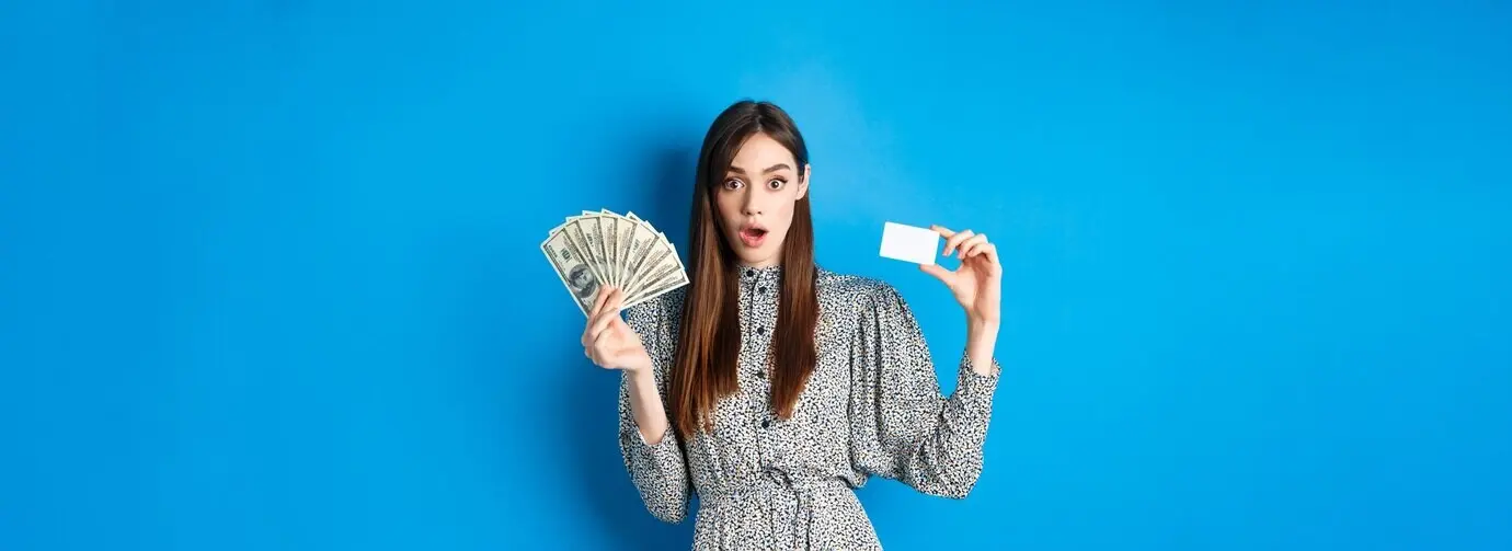Chica emocionada y afortunada mostrando una tarjeta de crédito de plástico y dinero en dólares, boquiabierta y asombrada, comprando algo.