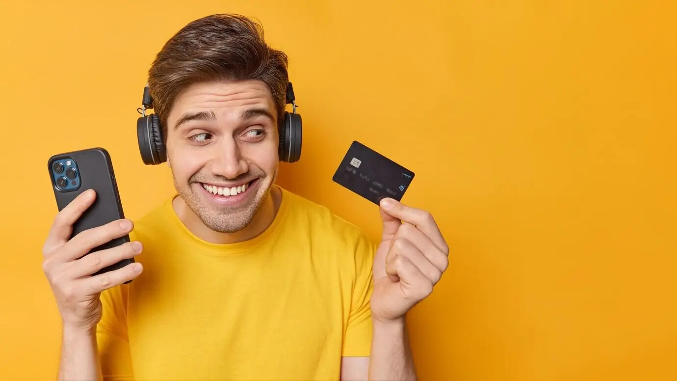Hombre guapo, optimista y despreocupado, sostiene un smartphone moderno y una tarjeta bancaria; usa una aplicación en el teléfono para realizar un pago en línea; aislado sobre fondo amarillo con espacio en blanco para tu contenido promocional.